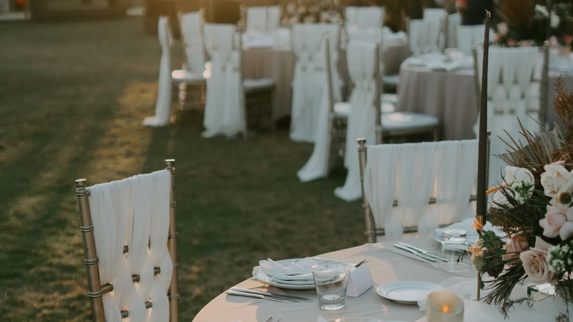 Outdoor wedding reception tables and dressed chairs laid out on grass at sunset