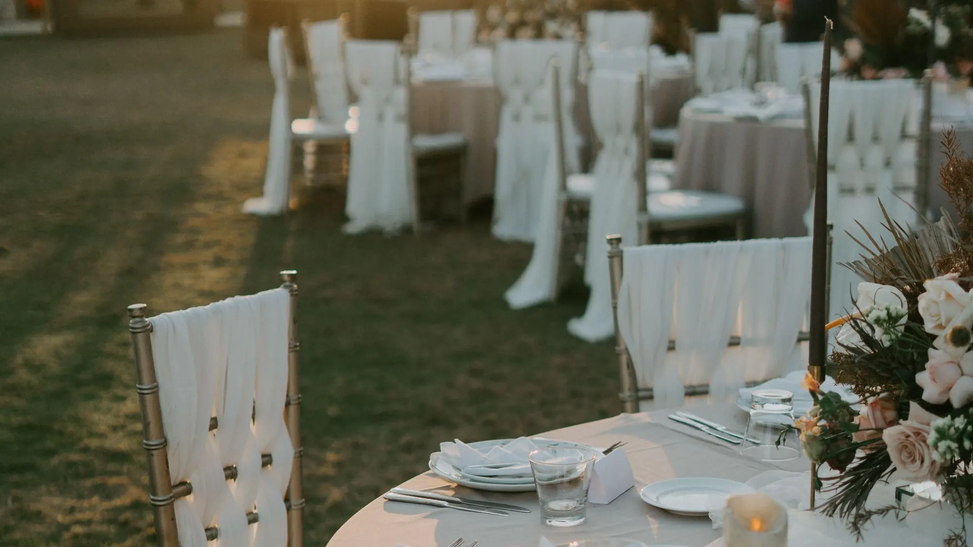 Outdoor wedding reception tables and dressed chairs laid out on grass at sunset